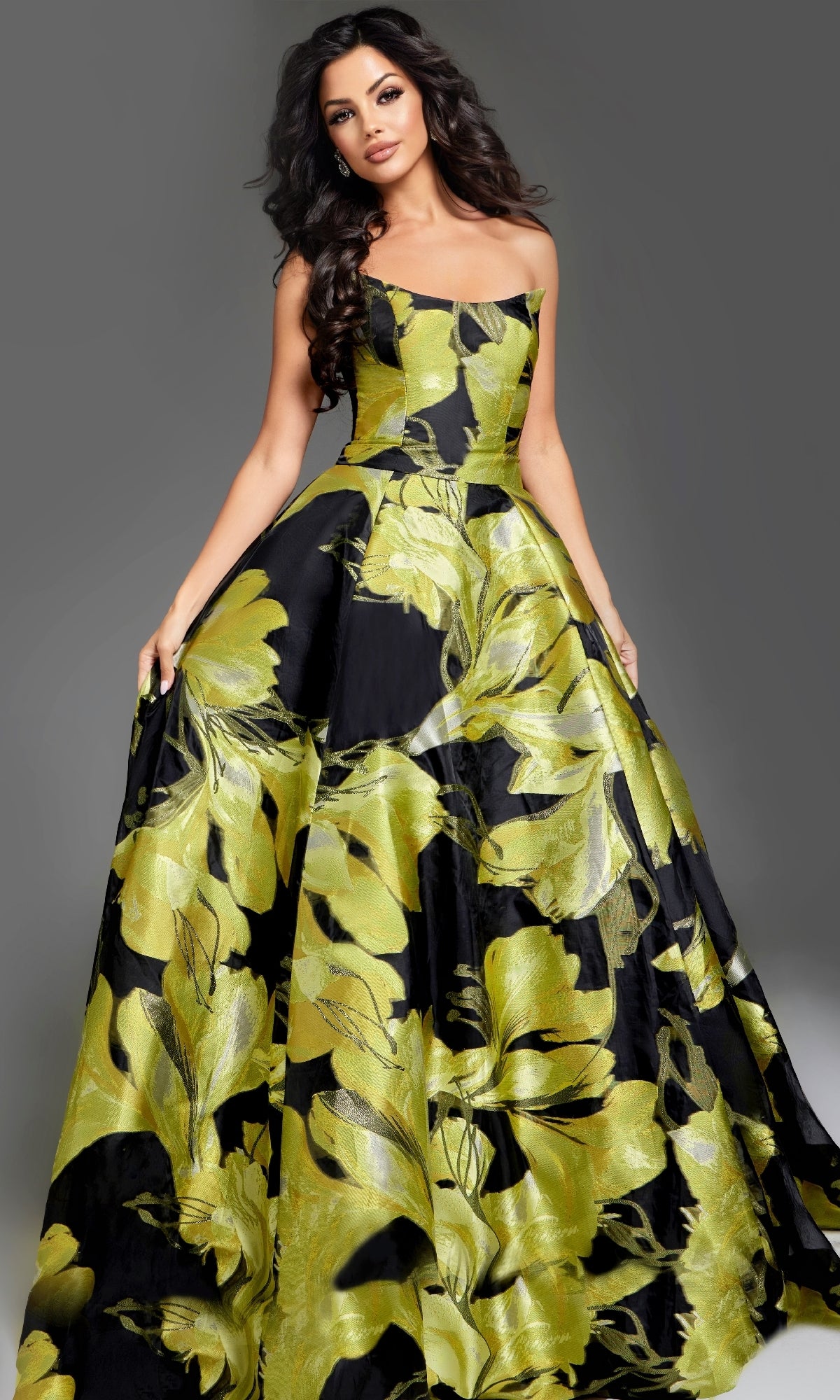 Woman wearing the Jovani 42435 Long Prom Dress, a strapless gown with a bold yellow-green floral print on black, posing against a plain grey backdrop.